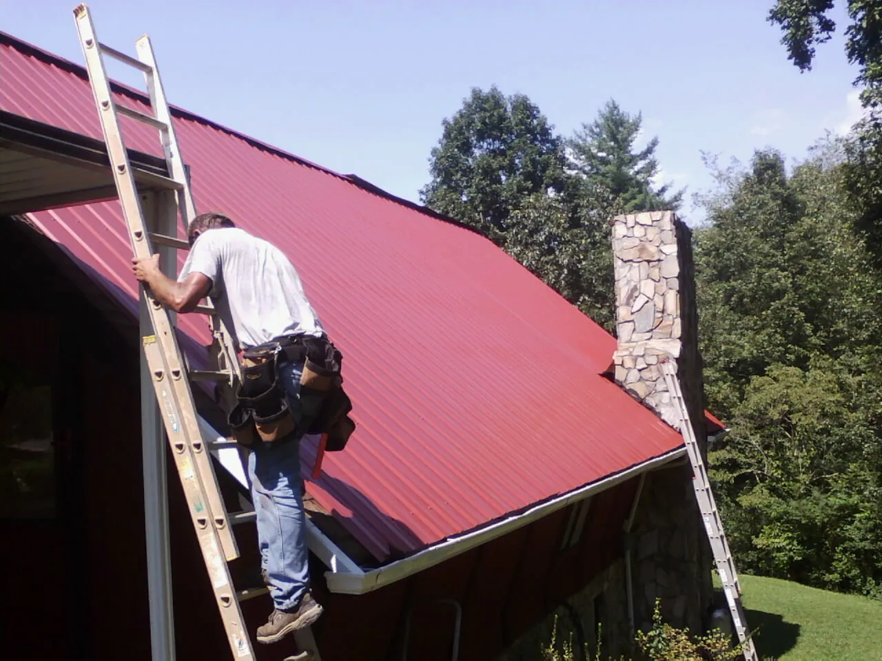 Roofer on a ladder beside a red metal roof and stone chimney — Soffit & Fascia Repair by Roofing Brothers in Glen Hope